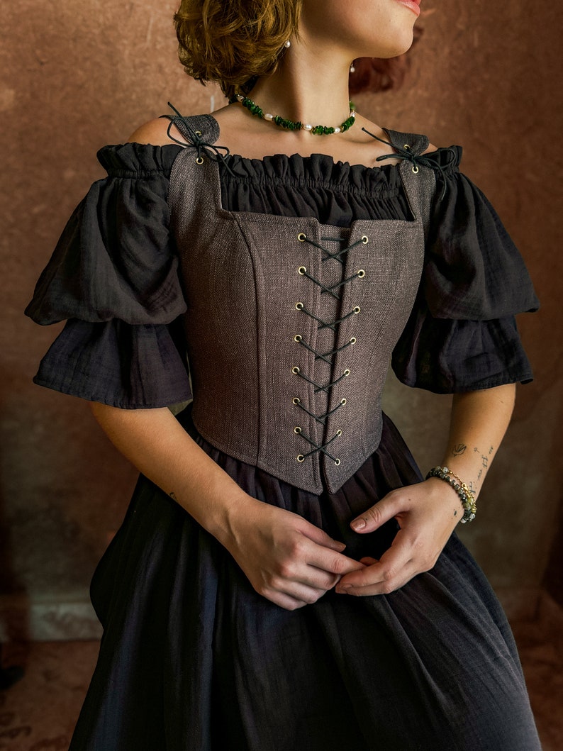 May include: A brown linen corset with black lacing and gold eyelets. The corset has a square neckline and is worn over a black off-the-shoulder top.