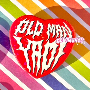 May include: Red heart-shaped pin with white text that reads "Old Man Trash" in a gothic font. The text is outlined in black. The pin has a glittery finish.