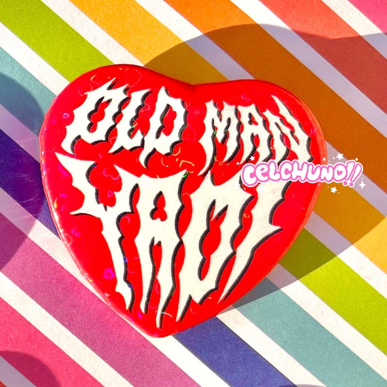 May include: Red heart-shaped pin with white text that reads "Old Man Trash" in a gothic font. The text is outlined in black. The pin has a glittery finish.