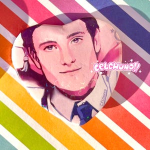 May include: A heart-shaped resin pin with a portrait of a person with dark hair and blue eyes. The pin has a pink background with glitter and the text "celchuno!" in white.