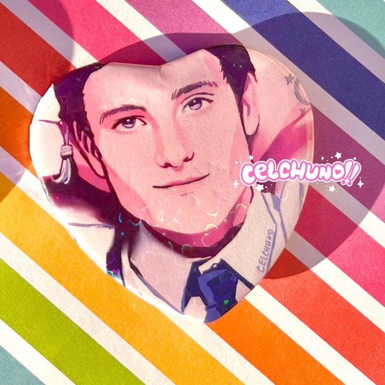 May include: A heart-shaped resin pin with a portrait of a person with dark hair and blue eyes. The pin has a pink background with glitter and the text "celchuno!" in white.