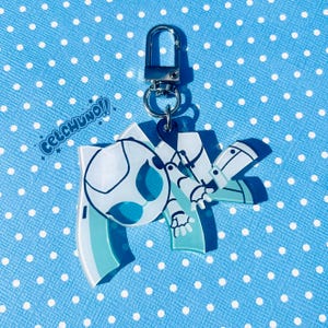 Puede incluir: Un llavero acr&iacute;lico azul y blanco con un dise&ntilde;o de dibujos animados y un cierre plateado. El llavero presenta un personaje estilizado con un bal&oacute;n de f&uacute;tbol y el texto "CELCHUNO!!". El fondo es una superficie azul con lunares blancos.