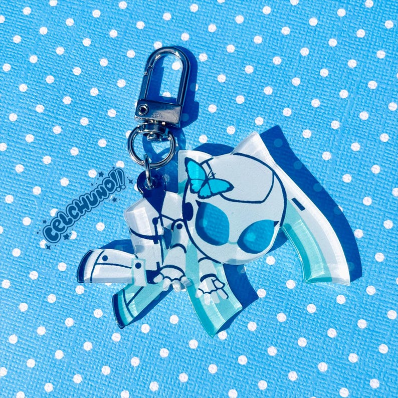 Puede incluir: Un llavero acr&iacute;lico azul y blanco con un dise&ntilde;o estilizado, una mariposa y el texto "CELCHUNO!!". Tiene un cierre y un anillo plateados. El fondo es una tela azul claro con lunares blancos.