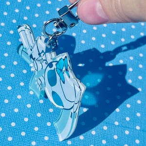 Puede incluir: Un llavero de acr&iacute;lico transparente con un dise&ntilde;o azul y blanco, sujeto a un cierre met&aacute;lico plateado. El llavero presenta un patr&oacute;n floral o abstracto estilizado. El fondo es una superficie azul claro con lunares blancos.