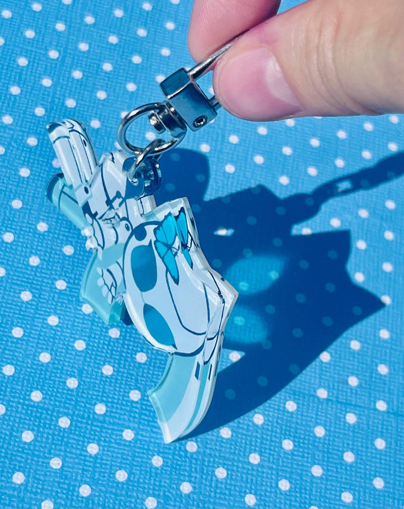 Puede incluir: Un llavero de acr&iacute;lico transparente con un dise&ntilde;o azul y blanco, sujeto a un cierre met&aacute;lico plateado. El llavero presenta un patr&oacute;n floral o abstracto estilizado. El fondo es una superficie azul claro con lunares blancos.