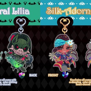 May include: Two double-sided acrylic keychains featuring anime characters. The first keychain features General Lilia, a green-skinned character with bat wings and a sword. The second keychain features Silk-Adorned Jamil, a brown-skinned character with blue and gold clothing and a flower in their hair. Both keychains come with a dangling color-shifting heart charm.