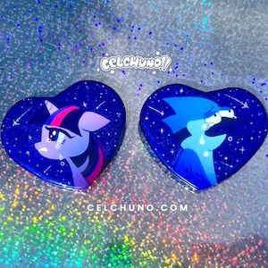 May include: Two heart-shaped pins featuring cartoon characters against a starry blue background. One pin depicts a purple pony with a pink mane and tears, the other a blue character. The text "CELCHUNO!!" and "CELCHUNO.COM" are visible.
