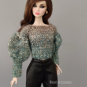 May include: A doll wearing a green and brown knitted sweater with large puffed sleeves and black trousers. The doll has long brown hair and is wearing gold earrings. The text "@HEART.OF.FLESH.DOLLS" is visible in the bottom right corner of the image.