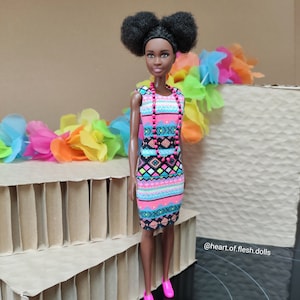 May include: A Black Barbie doll with a pink and blue geometric print dress and a pink beaded necklace. The doll has brown eyes, dark brown hair in two high buns, and is wearing pink shoes. The doll is standing on a cardboard background with a rainbow colored garland behind her.