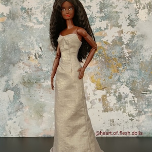 Könnte beinhalten: Eine schwarze Puppe, die ein langes, fließendes, goldfarbenes Kleid mit einem Rüschen-Saum trägt. Die Puppe hat langes, dunkles Haar und trägt roten Lippenstift. Die Puppe steht auf einem schwarzen Sockel vor einem strukturierten, blau-braunen Hintergrund. Der Text "@heart.of.flesh.dolls" ist in der unteren rechten Ecke des Bildes sichtbar.