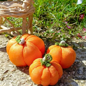 Puede incluir: Tres calabazas de tela naranja hechas a mano con detalles verdes y blancos descansan sobre una superficie de piedra. Las calabazas son de diferentes tamaños, con una pequeña silla de mimbre en el fondo. Decoración de otoño.