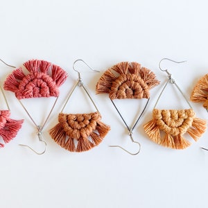 May include: Three pairs of handmade macrame earrings with a geometric design. The earrings are made with a combination of brown and pink yarn and feature a fringe detail. The earrings are attached to silver triangle-shaped hoops.