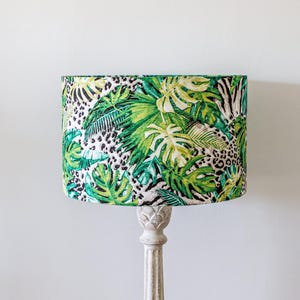 Può includere: Un paralume verde e bianco con un motivo a foglie tropicali. Il paralume è realizzato in tessuto e ha una forma cilindrica.