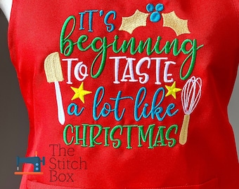 Könnte beinhalten: Rote Weihnachts-Schürze mit dem gestickten Text "It's beginning to taste a lot like Christmas". Der Text ist in Grün, Weiß und Blau mit gelben Sternen und Stechpalmenblättern. Es gibt auch gestickte Bilder eines Schneebesens und eines Spatels.