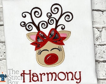 May include: White t-shirt with a red and brown embroidered reindeer with a red bow. The text "Harmony" is embroidered in red below the reindeer.