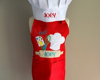 May include: Red chef apron with a white chef hat. The apron has a pocket and a design of a chef hat, rolling pin, and whisk. The hat and apron are personalized with the name "Joey".