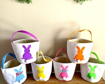 May include: Six Easter baskets with bunny silhouettes in different colors. The baskets are white with a white pom-pom tail on the bunny. The baskets are personalized with the names "Jhase", "Kyzer", "Kyrie", "Nova", and two other names.