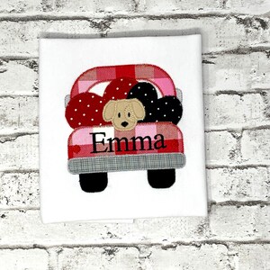 May include: A red and pink plaid truck with black wheels and a white background. The truck is carrying a dog and hearts. The name "Emma" is embroidered on the truck.