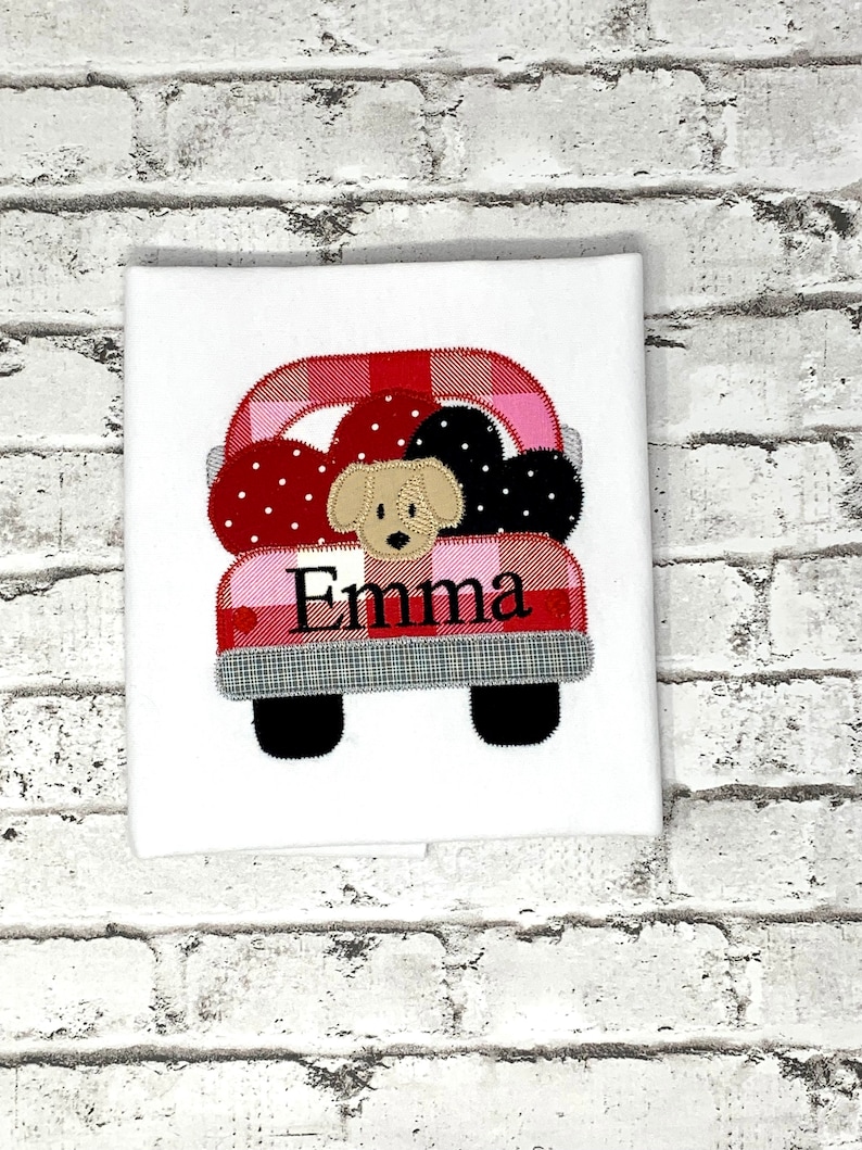 May include: A red and pink plaid truck with black wheels and a white background. The truck is carrying a dog and hearts. The name "Emma" is embroidered on the truck.