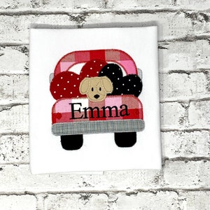 May include: A white t-shirt with a red and pink plaid truck with a brown dog in the bed. The truck has black wheels and the name "Emma" is embroidered on the back.