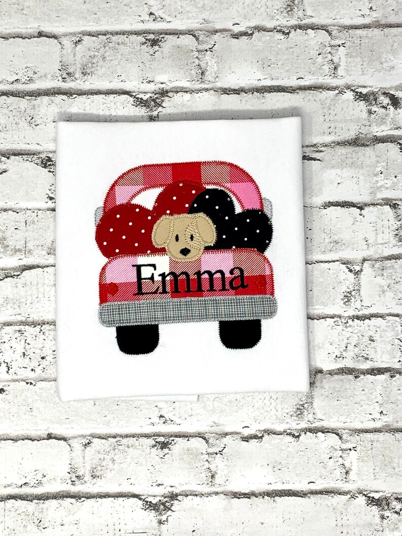 May include: A white t-shirt with a red and pink plaid truck with a brown dog in the bed. The truck has black wheels and the name "Emma" is embroidered on the back.