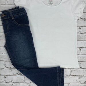 May include: A white short-sleeved t-shirt with a puff sleeve and a pink tag that says "AJ Blanks 4T" on a white brick background. A pair of dark blue denim jeans are lying on top of the shirt.
