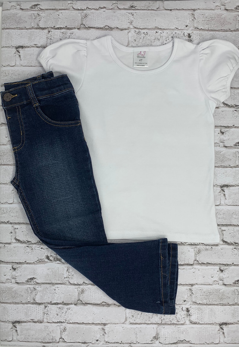 May include: A white short-sleeved t-shirt with a puff sleeve and a pink tag that says "AJ Blanks 4T" on a white brick background. A pair of dark blue denim jeans are lying on top of the shirt.