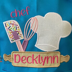 Pu&ograve; includere: Un grembiule blu turchese con un cappello da chef bianco, una frusta rosa e viola, una spatola con codette colorate e un mattarello con il nome "Decklynn" ricamato in rosa su uno sfondo beige. Il testo "chef" &egrave; ricamato in rosa sulla parte superiore del grembiule.