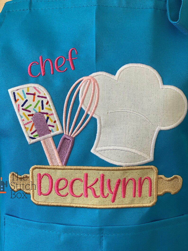 Pu&ograve; includere: Un grembiule blu turchese con un cappello da chef bianco, una frusta rosa e viola, una spatola con codette colorate e un mattarello con il nome "Decklynn" ricamato in rosa su uno sfondo beige. Il testo "chef" &egrave; ricamato in rosa sulla parte superiore del grembiule.