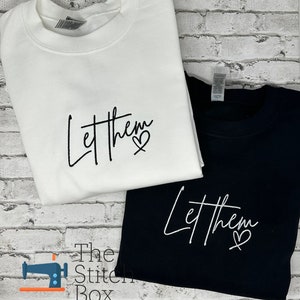 May include: Two sweatshirts, one white and one black, with the text "Let them" and a heart symbol embroidered in white on the black sweatshirt and black on the white sweatshirt.