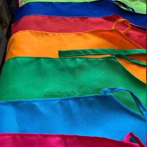Pu&ograve; includere: Una pila di grembiuli colorati in diverse tonalit&agrave; di rosa, verde, blu, rosso, arancione e giallo. I grembiuli sono realizzati in un tessuto liscio e tessuto e hanno bretelle regolabili.