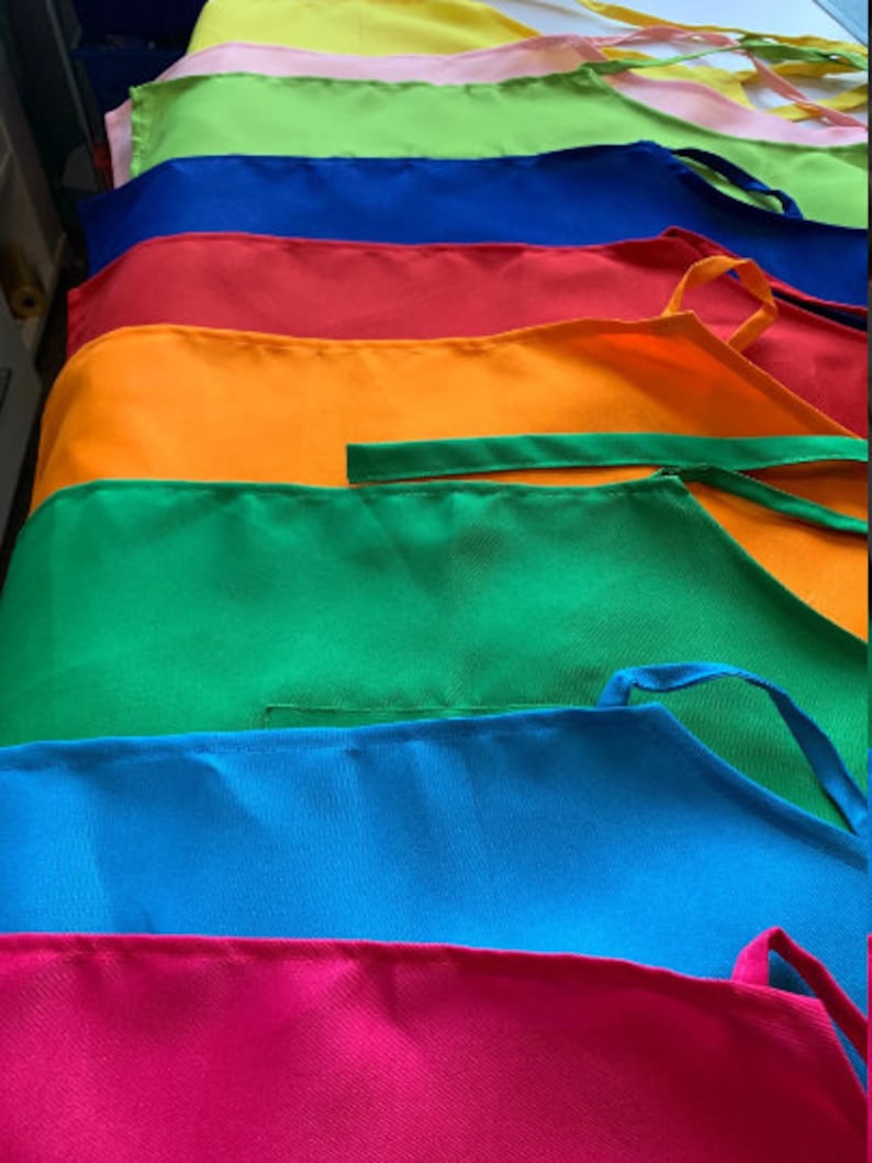 Pu&ograve; includere: Una pila di grembiuli colorati in diverse tonalit&agrave; di rosa, verde, blu, rosso, arancione e giallo. I grembiuli sono realizzati in un tessuto liscio e tessuto e hanno bretelle regolabili.