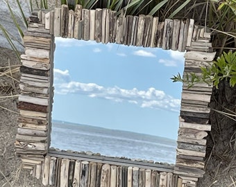 May include: A square mirror with a frame made of driftwood. The mirror reflects a blue sky with white clouds and a body of water.