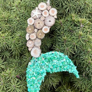 May include: A handcrafted mermaid ornament with a tail made of small, vibrant green stones. The mermaid's body is composed of various wooden slices and a starfish, suspended by a natural twine string. The ornament is set against a backdrop of green foliage.