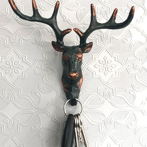 May include: A decorative wall-mounted key holder shaped like a deer head with antlers. The deer head is dark with copper-coloured accents, and a key ring holds a set of keys. The holder is mounted on a white textured wall.