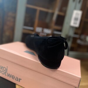 May include: Black suede ballet flat with a bow detail on the heel, on a pink "koi footwear" box. The shoe has a rounded toe and a low heel. The box is light pink with the brand name printed in a dark grey font.