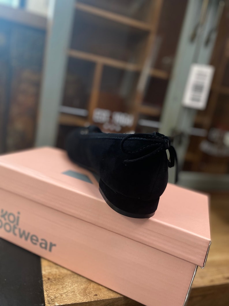 May include: Black suede ballet flat with a bow detail on the heel, on a pink "koi footwear" box. The shoe has a rounded toe and a low heel. The box is light pink with the brand name printed in a dark grey font.