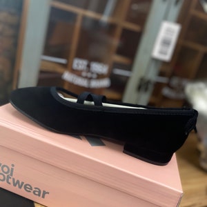 May include: A black ballet flat with a rounded toe and a small heel, featuring a strap across the top. The shoe is displayed on a pink box with the brand name "Koi Footwear" printed on the side. The shoe is made of a soft material.