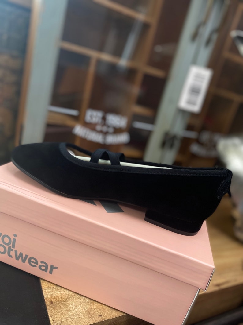 May include: A black ballet flat with a rounded toe and a small heel, featuring a strap across the top. The shoe is displayed on a pink box with the brand name "Koi Footwear" printed on the side. The shoe is made of a soft material.