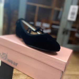 May include: Black velvet flats with a pointed toe and strap detail, on a pink shoe box. The shoe box has the word "wear" printed on it. The shoes are a classic style.