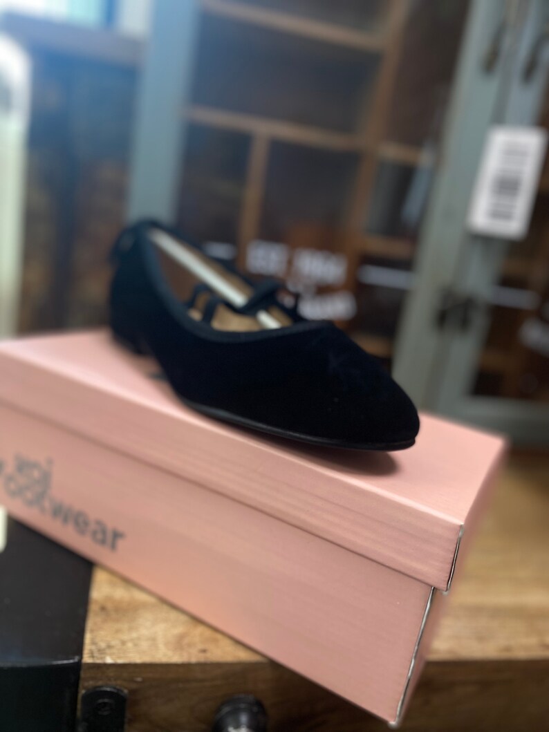 May include: Black velvet flats with a pointed toe and strap detail, on a pink shoe box. The shoe box has the word "wear" printed on it. The shoes are a classic style.