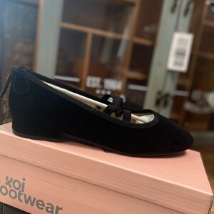 May include: Black ballet flats with a rounded toe and a small heel, featuring a strap design. The flats are displayed on a pink box with the text "koi footwear". The shoes appear to be made of suede or a similar material.