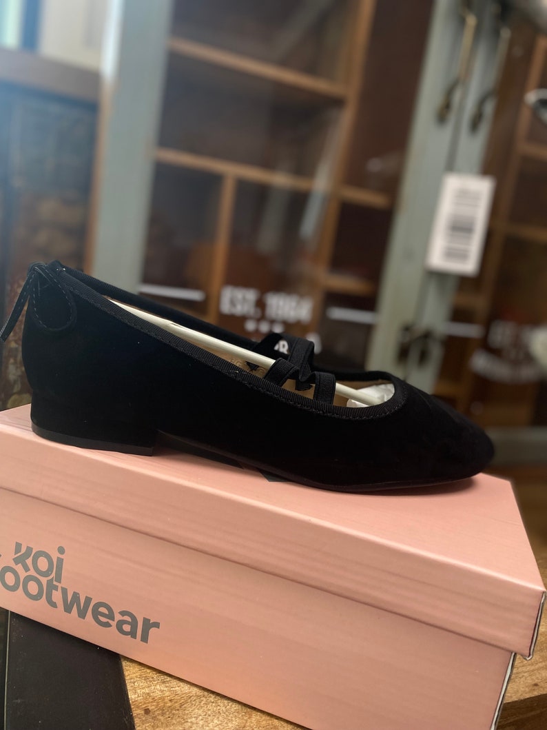 May include: Black ballet flats with a rounded toe and a small heel, featuring a strap design. The flats are displayed on a pink box with the text "koi footwear". The shoes appear to be made of suede or a similar material.