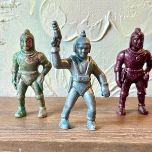 May include: Three vintage toy astronaut figures in metallic colors. The figures are in shades of green, silver, and burgundy. The silver astronaut is holding a ray gun. The figures are standing on a wooden surface.