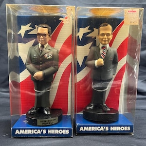 May include: Two boxed figurines of American heroes, General Colin Powell and President Ronald Reagan. The figurines are in a standing pose and are wearing their signature attire. The boxes are decorated with an American flag design and the words "America's Heroes".