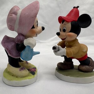 May include: Two ceramic figurines of cartoon characters. One is a female mouse wearing a pink bonnet and purple dress, holding a blue watering can. The other is a male mouse in a red hat and tan jacket, holding a small object. Both are on round bases.