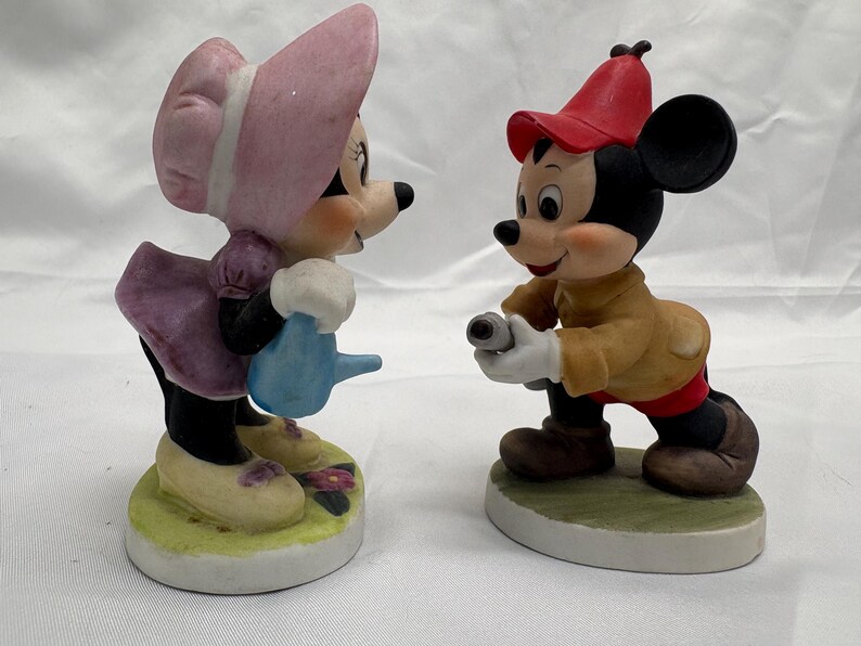 May include: Two ceramic figurines of cartoon characters. One is a female mouse wearing a pink bonnet and purple dress, holding a blue watering can. The other is a male mouse in a red hat and tan jacket, holding a small object. Both are on round bases.