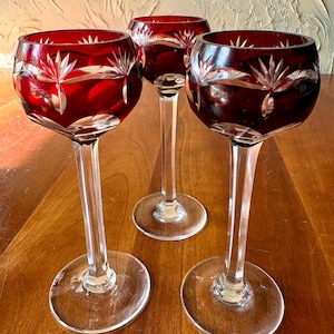 May include: Three ruby red wine glasses with clear stems and bases. The glasses have a cut glass design with a starburst pattern around the rim. The glasses are displayed on a wooden surface.