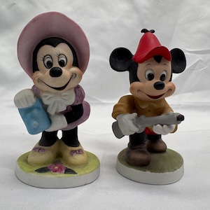 May include: Two ceramic figurines of cartoon characters. One is a female mouse wearing a pink hat and purple dress, holding a blue item. The other is a male mouse in a red hat and brown outfit, holding a gray object. Both are on circular bases.