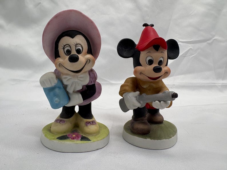 May include: Two ceramic figurines of cartoon characters. One is a female mouse wearing a pink hat and purple dress, holding a blue item. The other is a male mouse in a red hat and brown outfit, holding a gray object. Both are on circular bases.